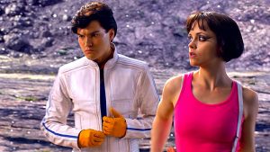 A man in a white jacket and orange gloves stands next to a woman in a bright pink tank top; both look in different directions outdoors.