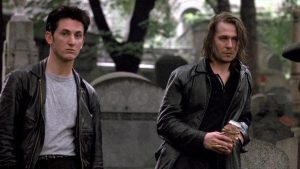 Two men wearing dark jackets stand in a cemetery; one holds a paper bag while gravestones and trees appear in the background.
