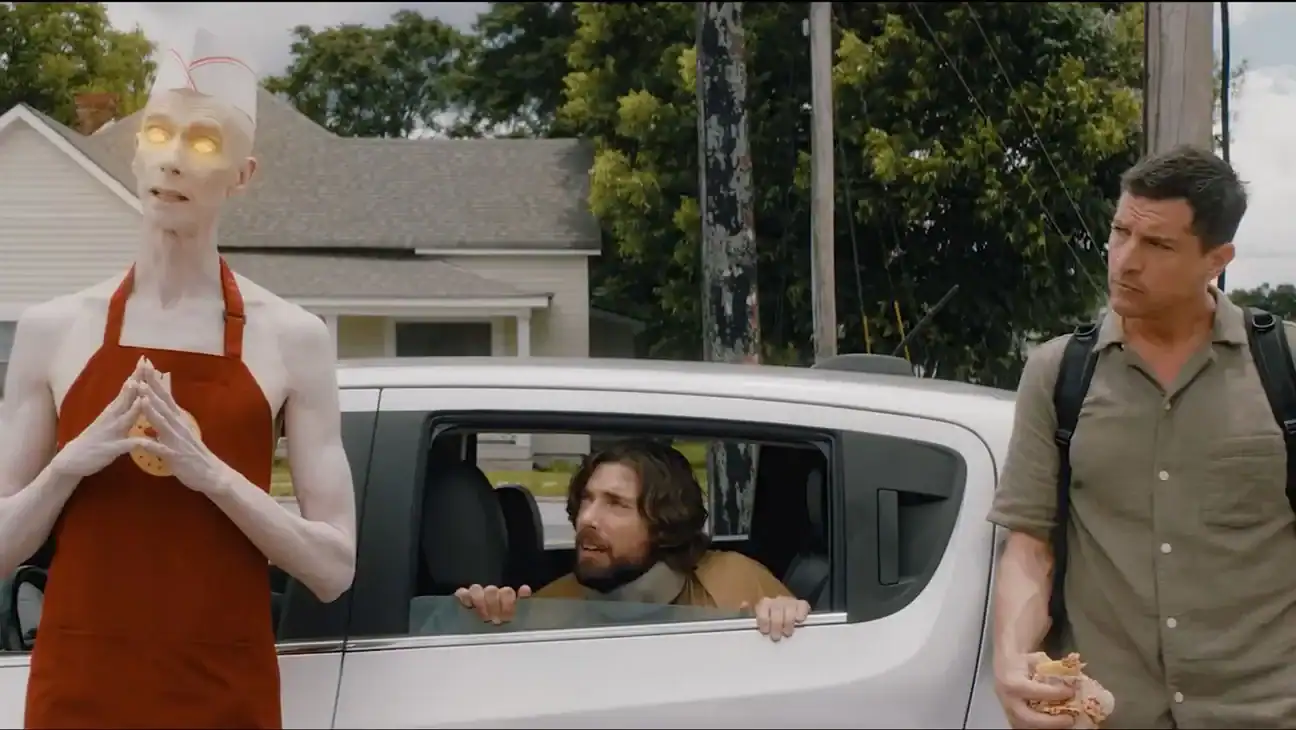 A tall, pale figure with glowing eyes stands by a car, while one man leans out of the car window and another stands nearby eating a sandwich.