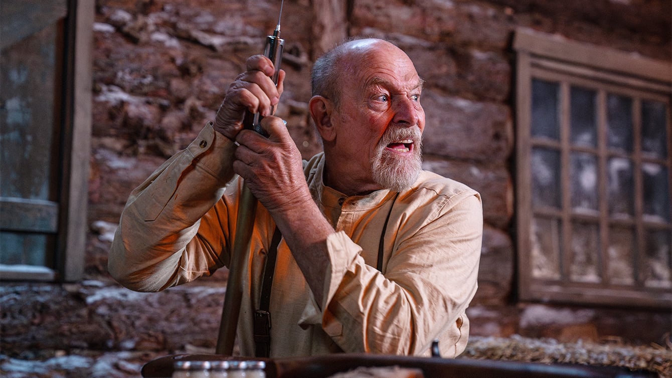 An older man with a bald head and gray beard holds a rifle and looks to the side inside a rustic wooden cabin with a window in the background.