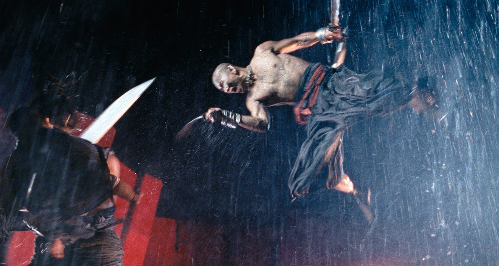 A shirtless man with swords leaps through heavy rain, facing an opponent with a raised blade in a dramatic action scene.