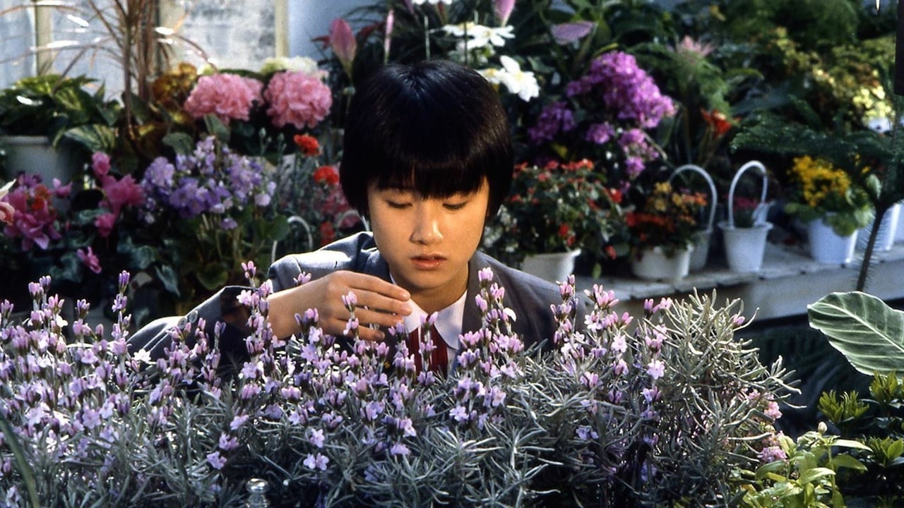 A person in a suit examines purple flowers in a garden or greenhouse filled with various potted plants and colorful blooms.