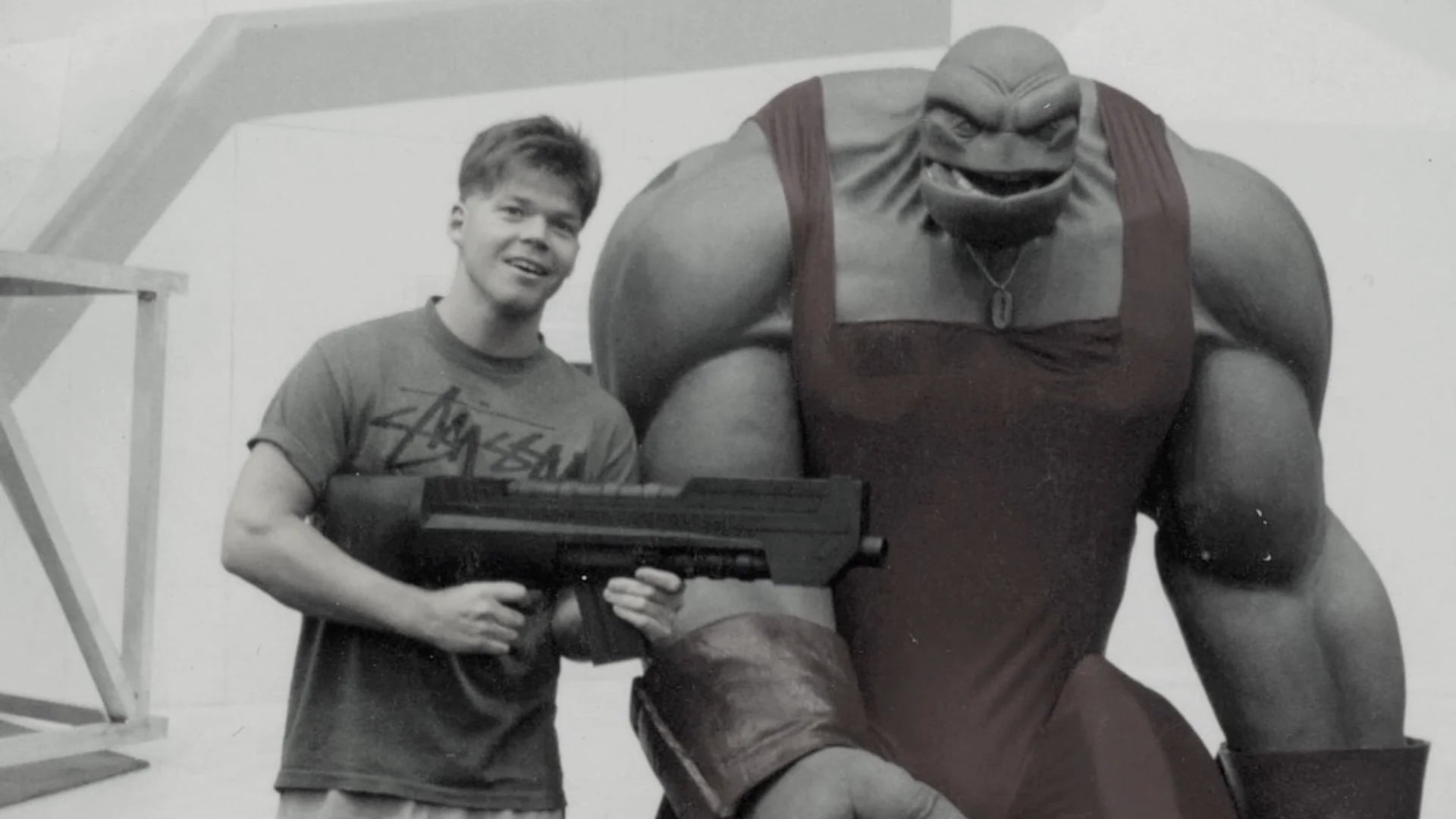 A young man holding a large prop gun stands next to a muscular humanoid creature in a one-piece outfit; the photo is in black and white.