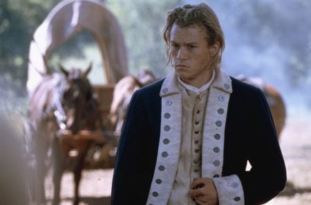 A young man in a historical military uniform stands in front of a horse-drawn covered wagon, holding his hand to his abdomen with a serious expression.