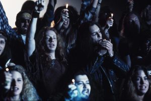 A crowd of people holds up lit lighters at an event. Some individuals have long hair and casual clothing, and one person in the foreground is lighting a cigarette.