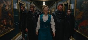 A woman in a blue dress leads four men in black cloaks down a hallway lined with paintings. All have serious expressions.