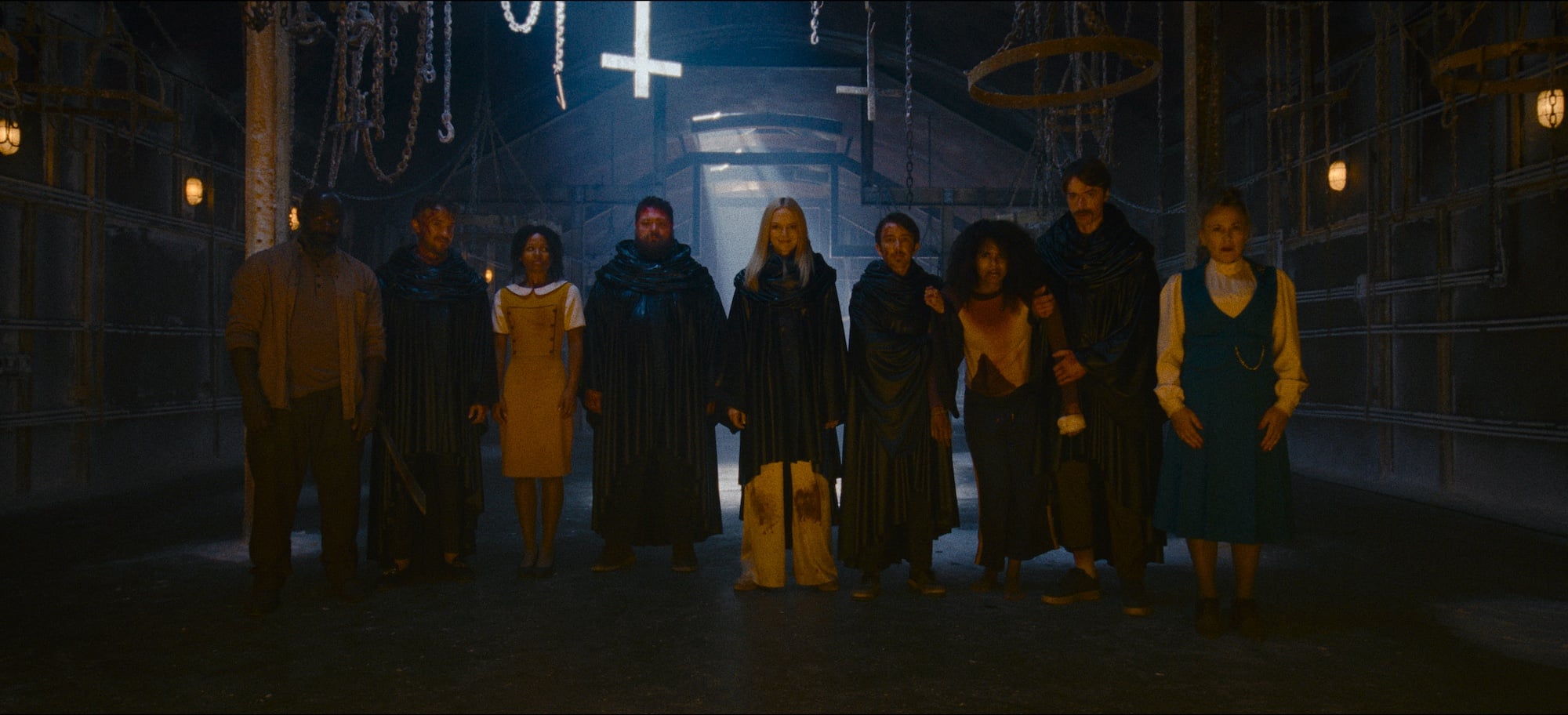 A group of people stand in a dimly lit room with hanging chains, circular frames, and an upside-down cross on the ceiling.
