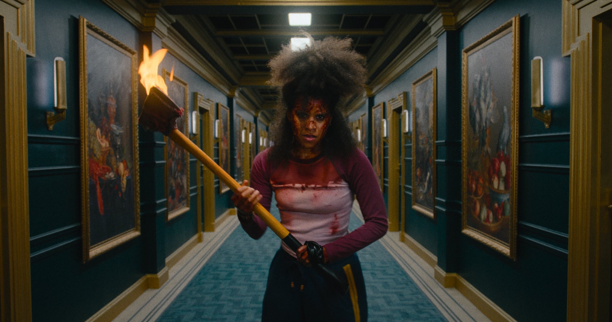 A person with disheveled hair and face paint, wearing a bloodstained shirt, stands in a hallway holding a torch with flames.