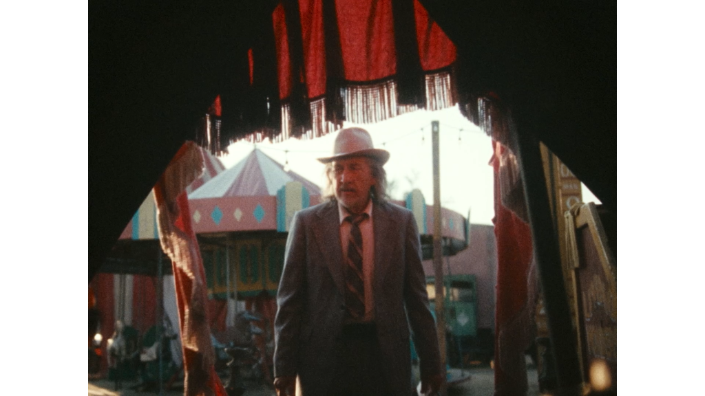 A man in a suit and hat stands outside a tent entrance, with carnival rides and colorful structures visible in the background.