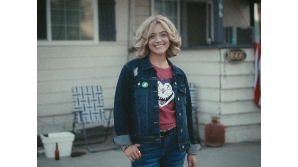 A person with shoulder-length blonde hair, wearing a denim jacket and jeans, stands smiling in front of a house with patio chairs and other outdoor items visible.
