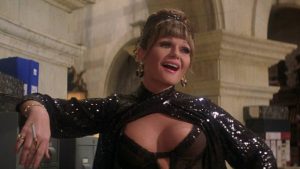 A woman in a sparkling black dress with jewelry poses indoors, smiling and holding a cigarette, with office shelves and architecture in the background.