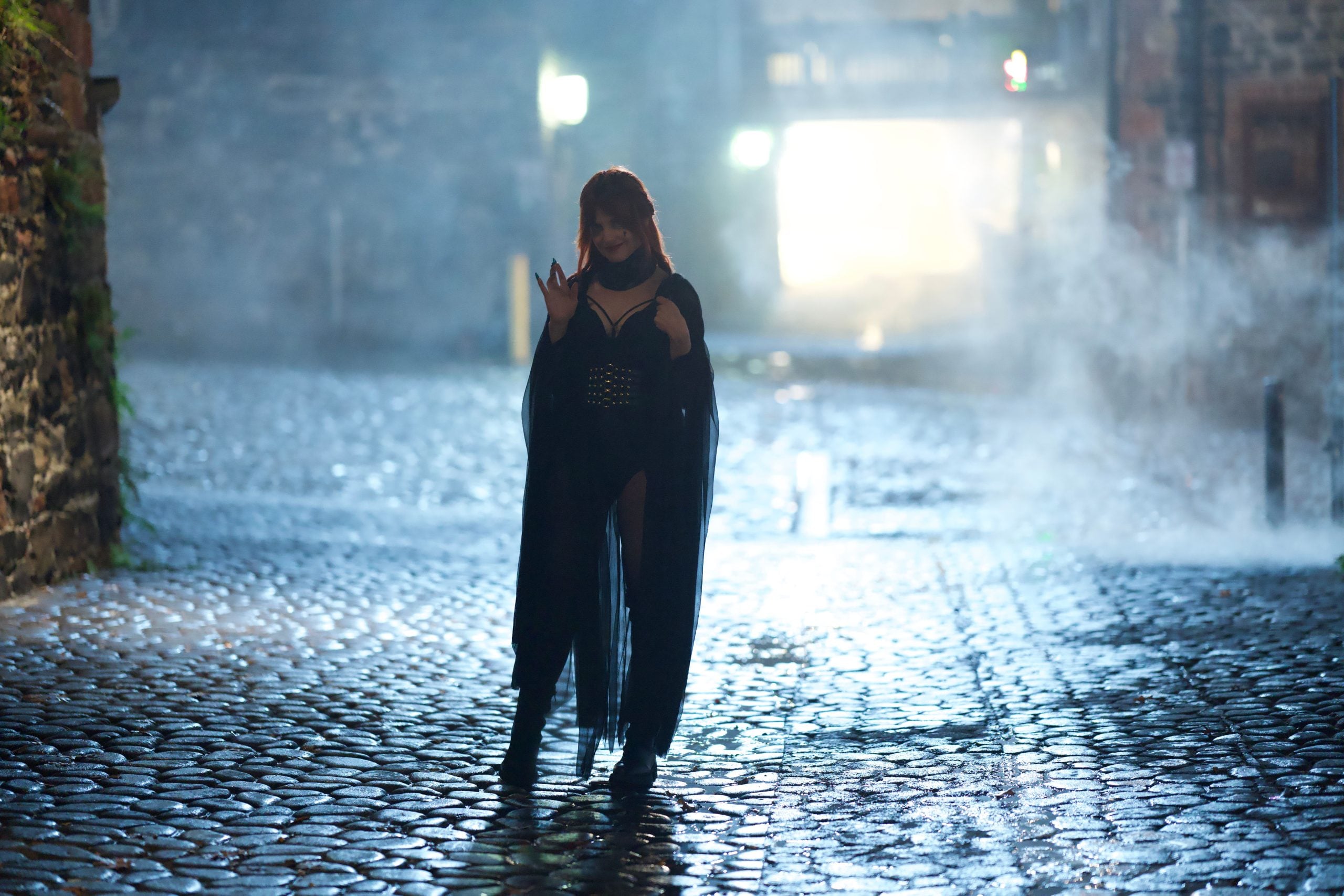 A person in a dark, dramatic costume stands on a wet cobblestone street at night, surrounded by mist and dim lighting.