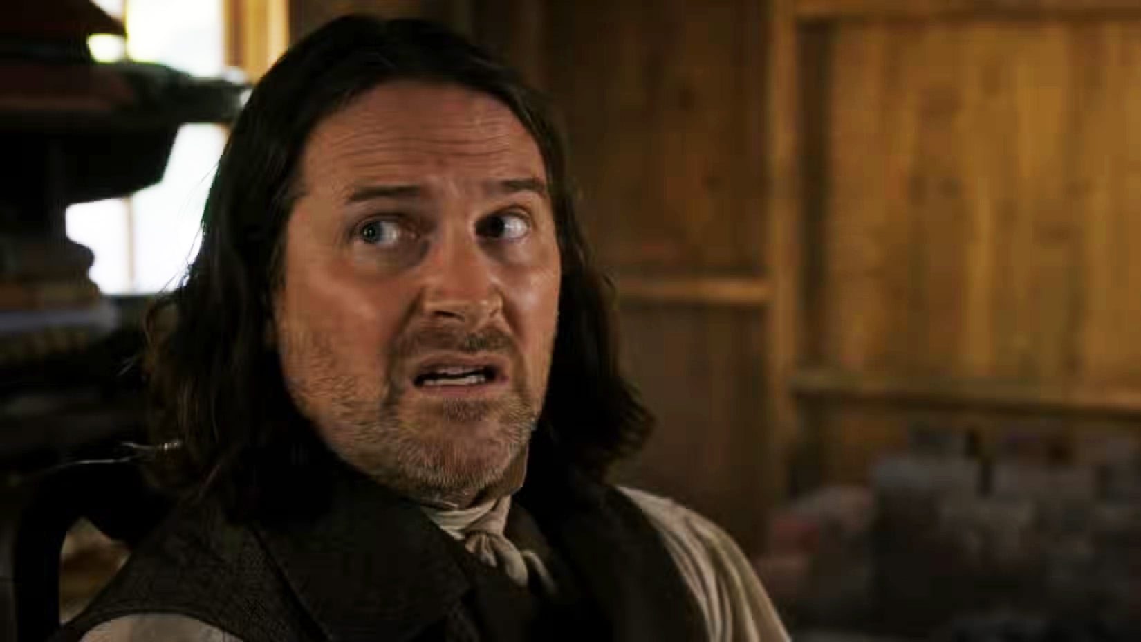 A man with shoulder-length hair and a beard looks to the side with a worried or uneasy expression, seated indoors in a wooden room.