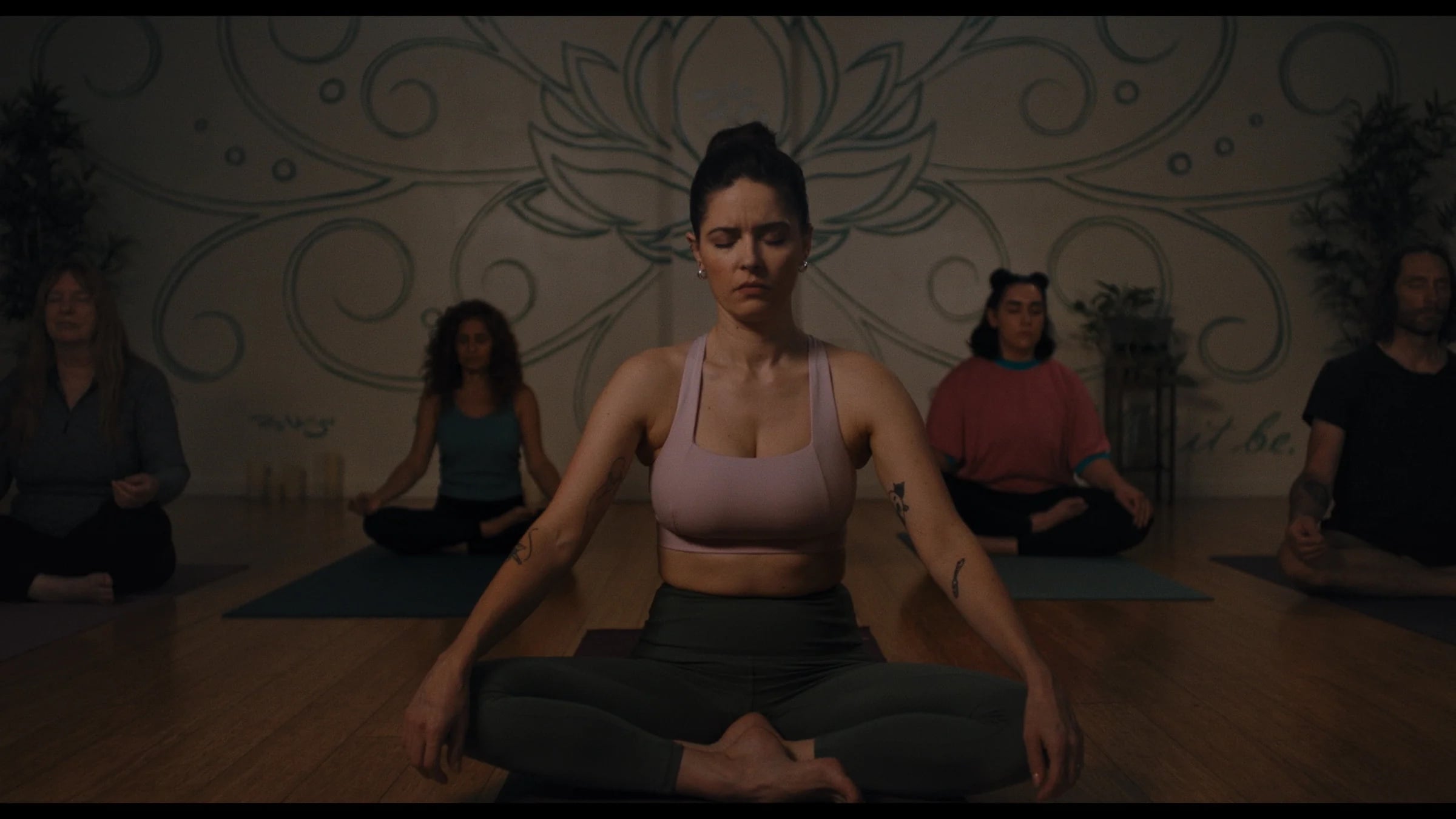 A group of people sit cross-legged on yoga mats in a dimly lit studio, eyes closed, meditating in front of a wall with a large, decorative mural.