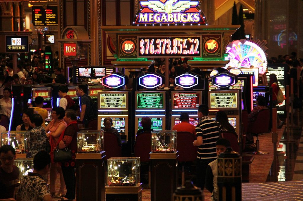 Crowd of people playing slot machines and table games in a busy, brightly lit casino with a large Megabucks jackpot sign overhead.