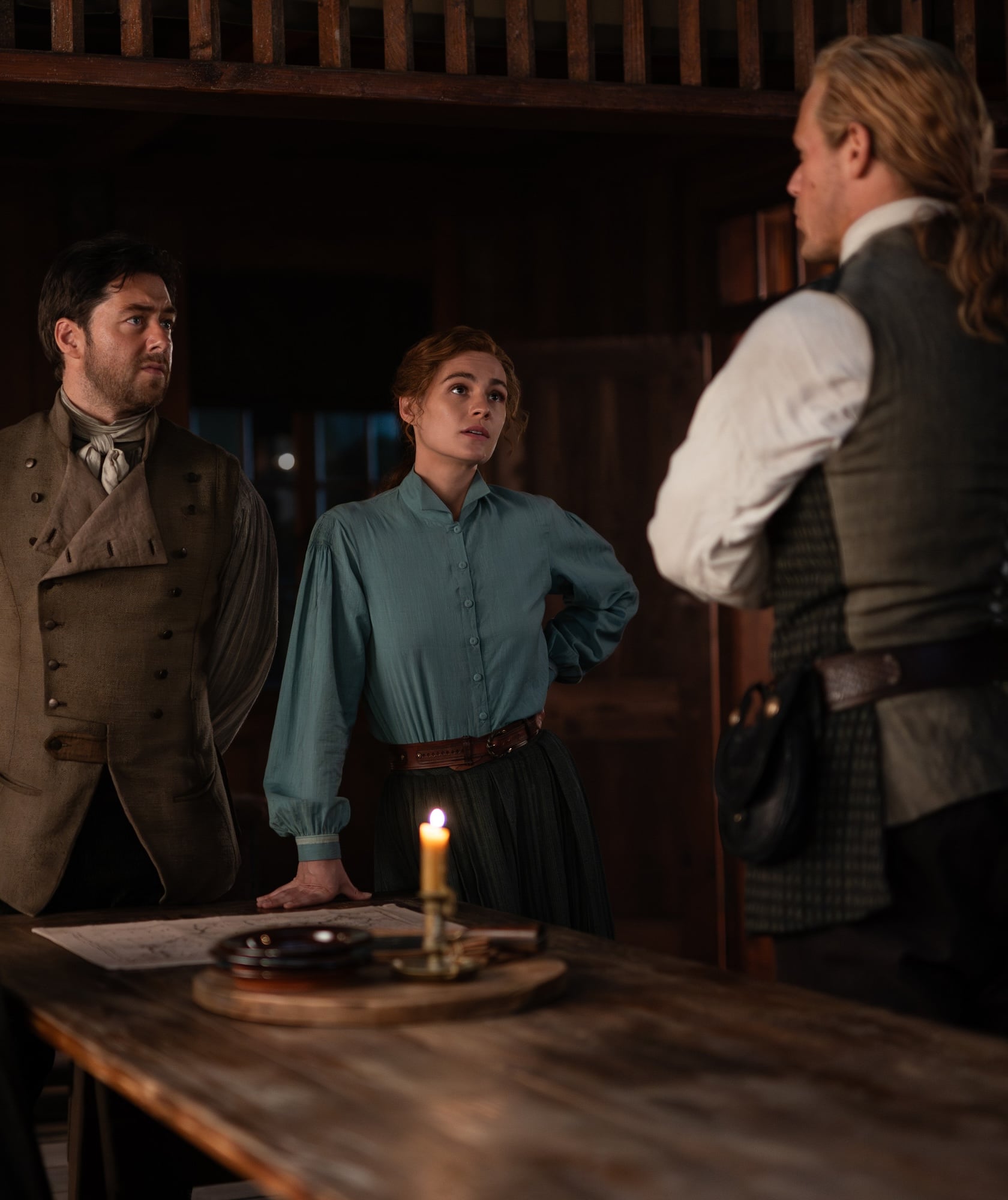 Three people in period clothing stand around a wooden table with a lit candle and papers, engaged in a serious conversation inside a dimly lit rustic room.