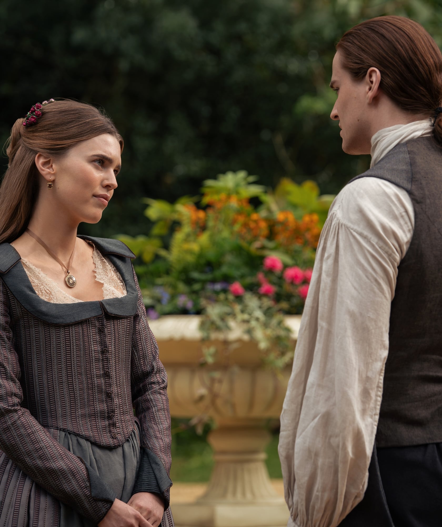 A woman and a man in historical clothing stand facing each other outdoors, with a large ornamental planter and greenery in the background.