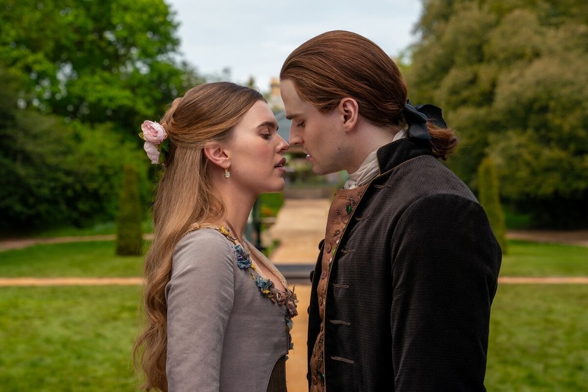 A woman and a man in historical costumes stand close together outdoors, about to kiss, with a garden path and greenery in the background.