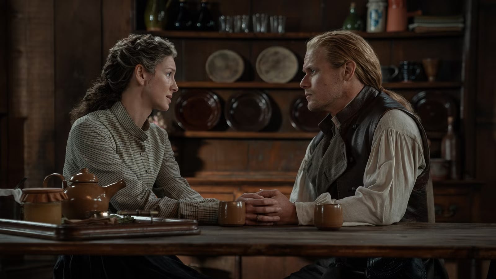A man and woman sit at a wooden table holding hands, looking at each other seriously in a rustic kitchen setting with dishes and cups in the background.