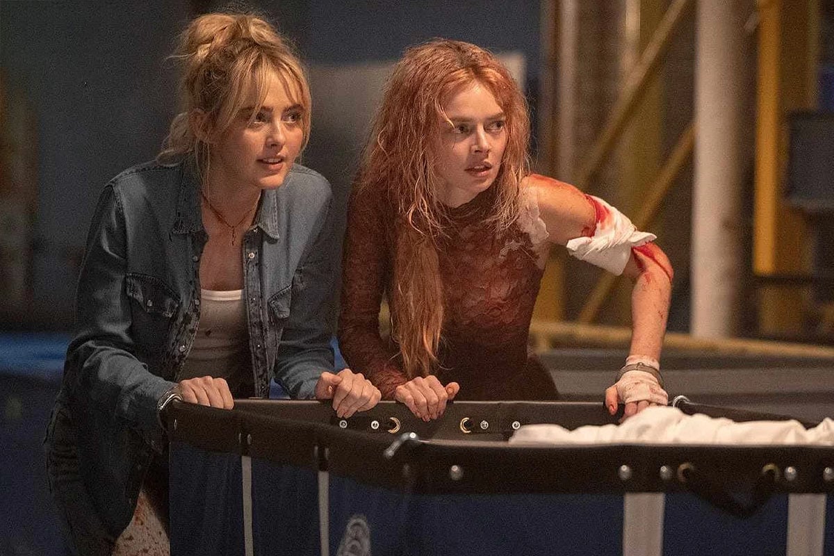 Two young women lean over a stretcher; one has messy hair and a bloody arm, both look intently ahead in a dimly lit industrial setting.