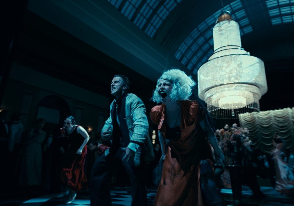 A man and a woman in dramatic poses move through a grand, dimly lit ballroom with a large chandelier overhead and people in the background.