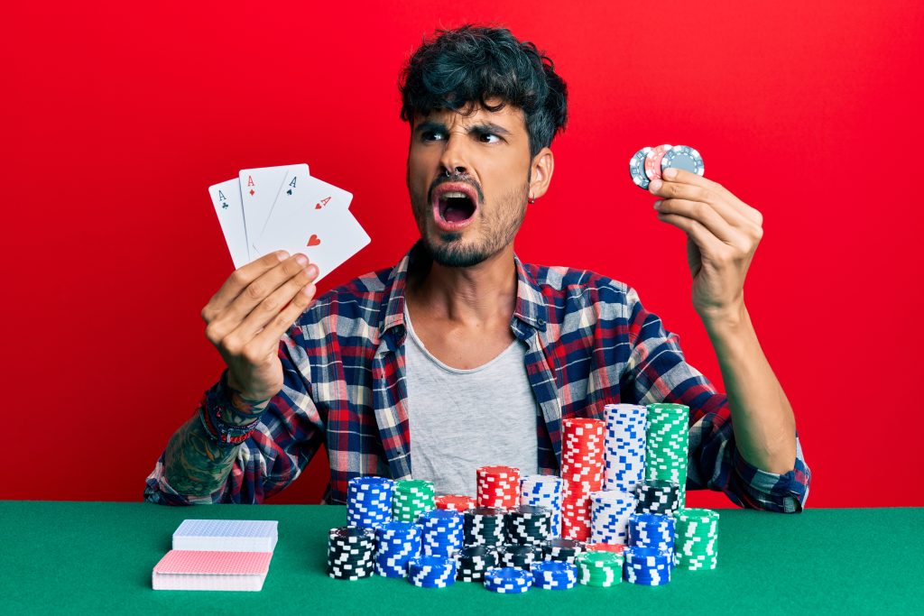A man sits at a poker table holding four aces in one hand and poker chips in the other, looking surprised. Stacks of chips and playing cards are in front of him against a red background.