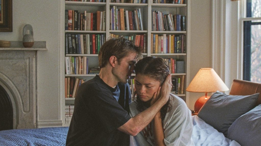 A man and woman sit on a bed in a bedroom, the man gently holding the woman's head as she looks down, with a bookshelf and lamp in the background.