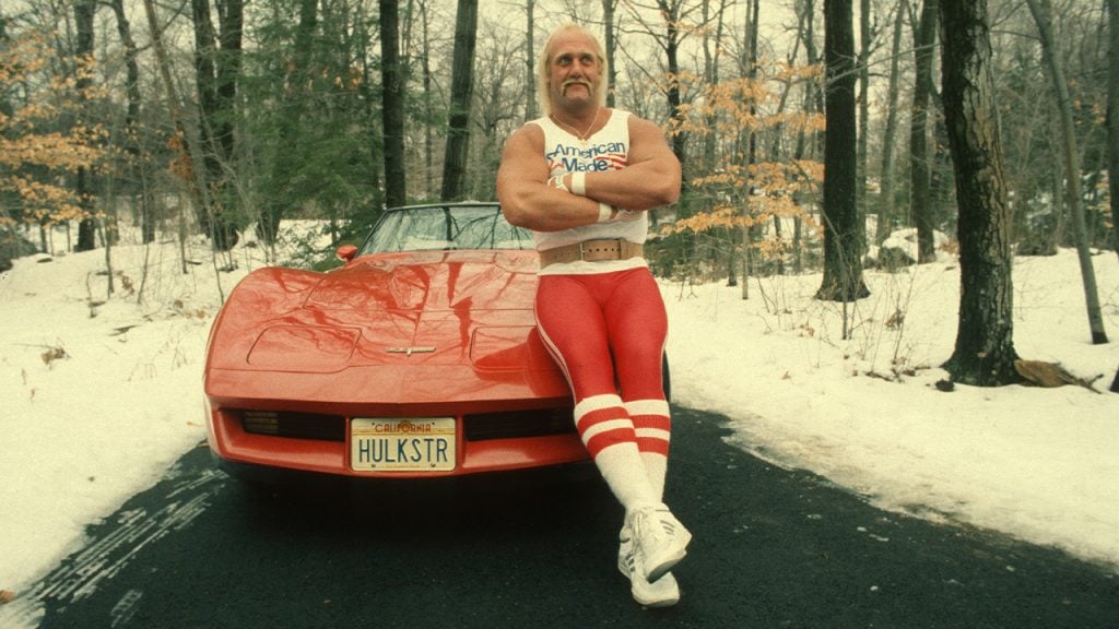 A muscular man in red and white workout clothes stands with arms crossed, leaning on a red Corvette with a "HULKSTR" license plate in a snowy, wooded area.