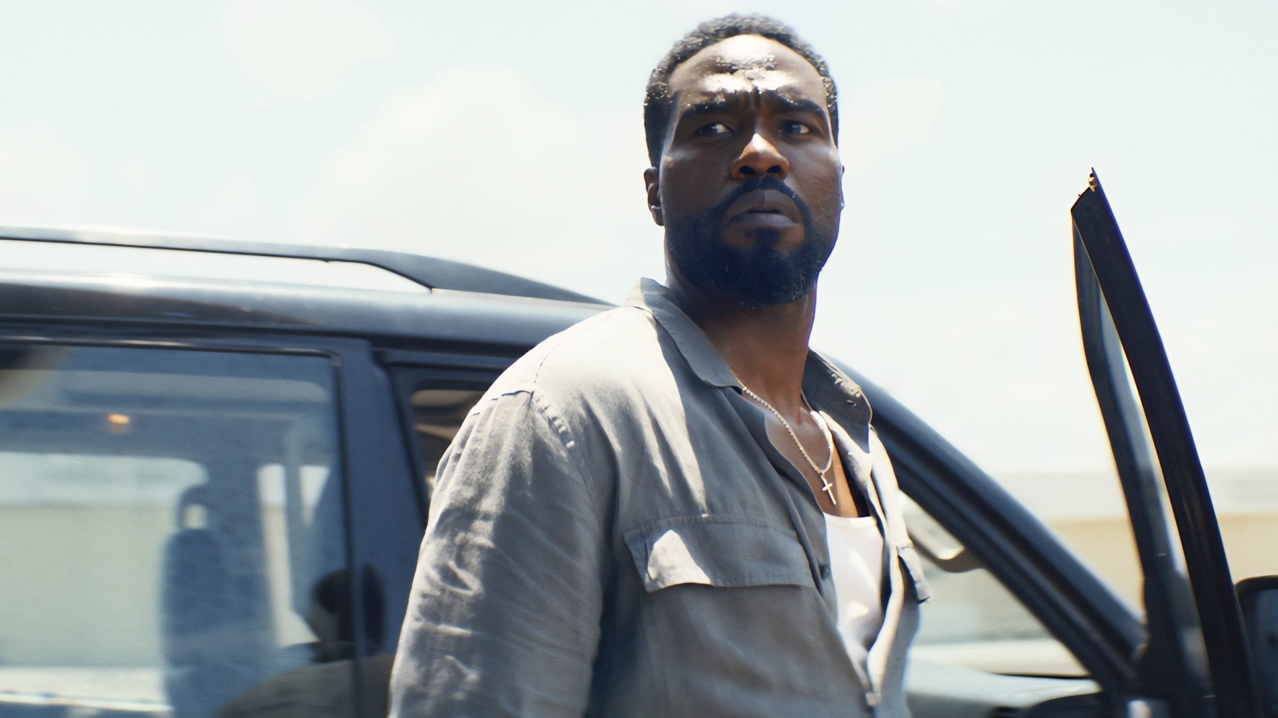 A man in a gray shirt stands beside an open car door, looking intently into the distance under bright daylight.