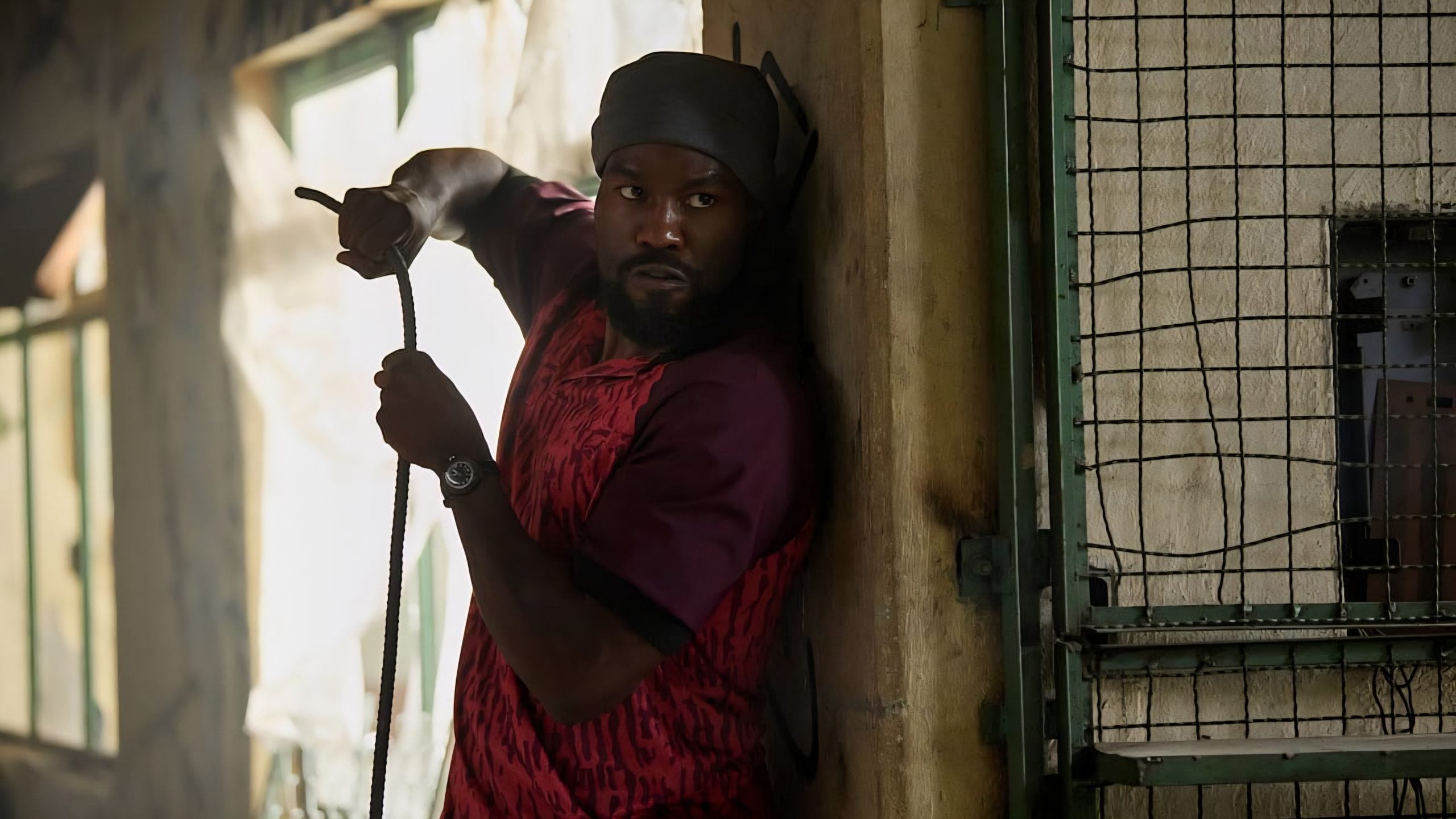 A man in a red shirt and headscarf stands alert against a wall, holding a rope and looking to his left, near a metal fenced gate in a dimly lit setting.
