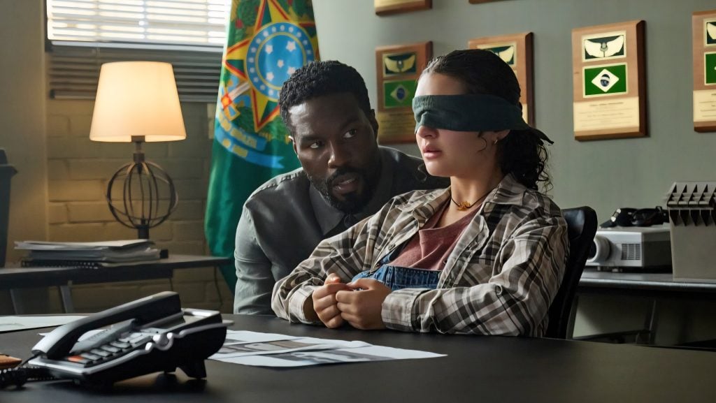 A man leans toward a blindfolded woman sitting at a desk with documents, a lamp, and plaques visible in the background.