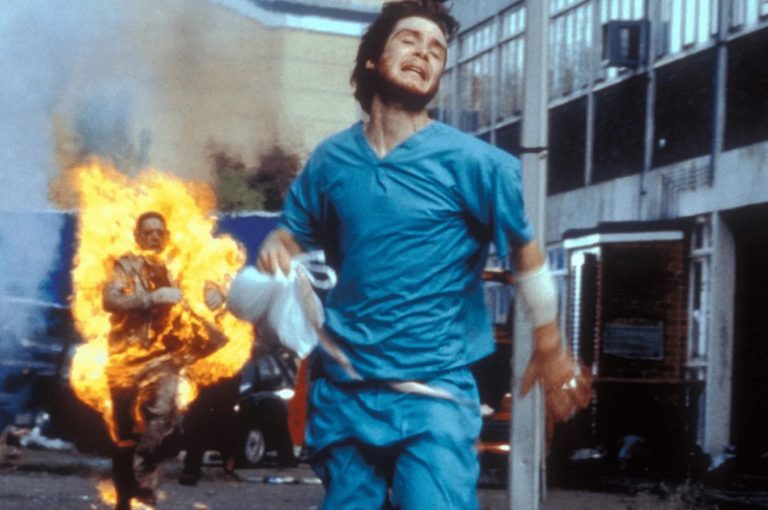 A man in blue scrubs runs through a street while another person, engulfed in flames, chases him from behind.