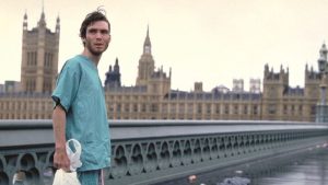 A man in hospital scrubs holding a plastic bag stands alone on an empty bridge with the Palace of Westminster in the background.