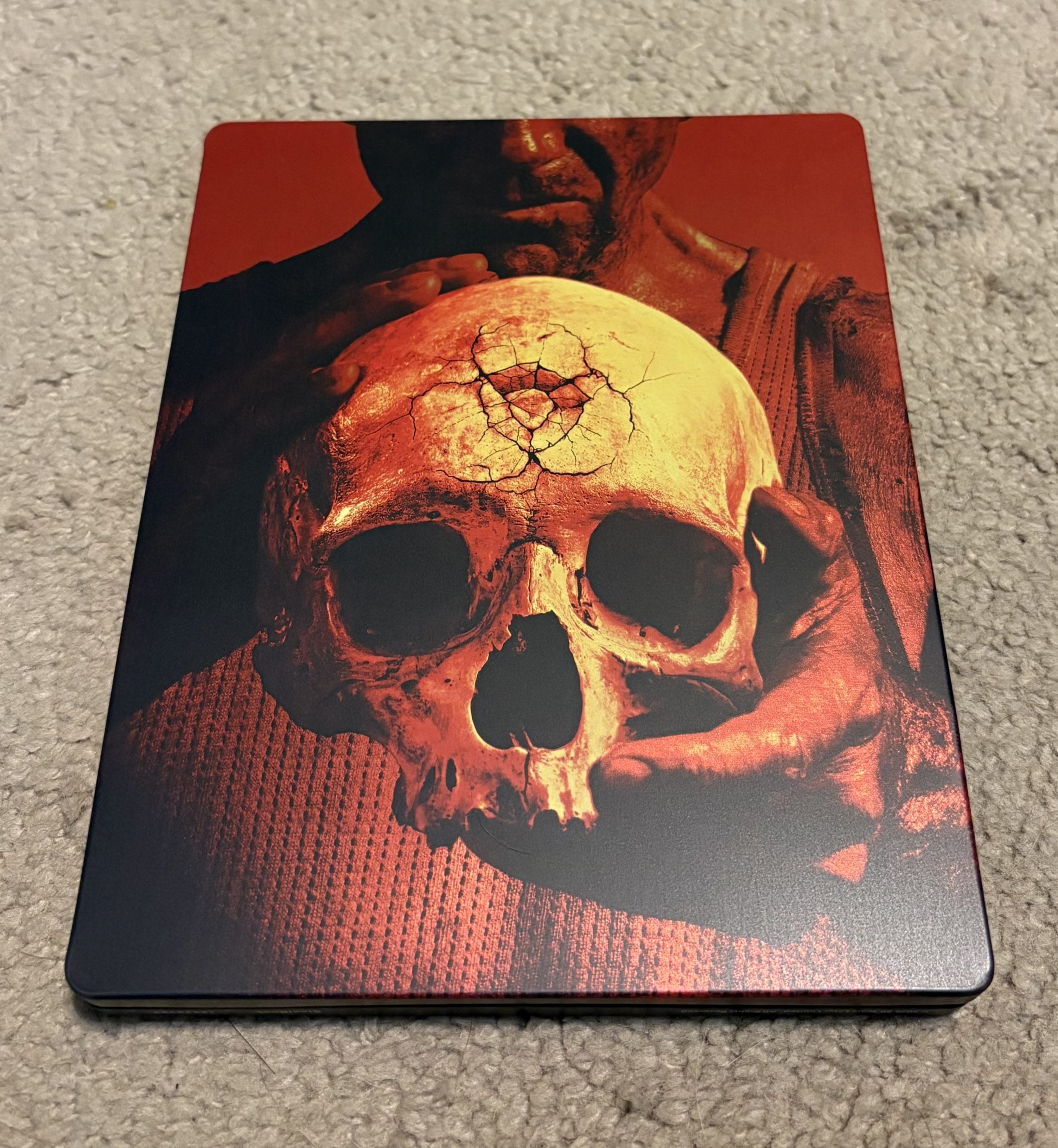 A person holding a cracked human skull with both hands; the background and lighting have a reddish tone.