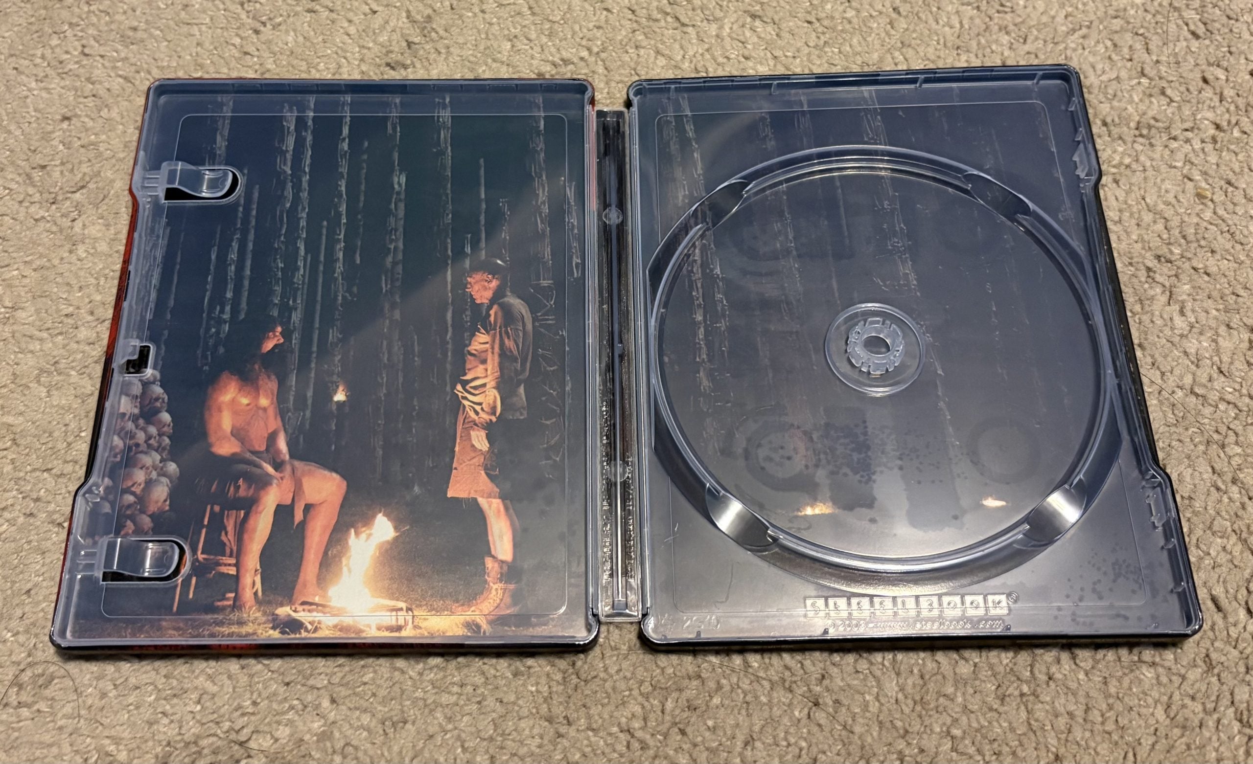 An open, empty Blu-ray case lying on carpet, with interior artwork showing two men by a fire, one seated and nude, surrounded by skulls, and the other standing, both in a forest setting.
