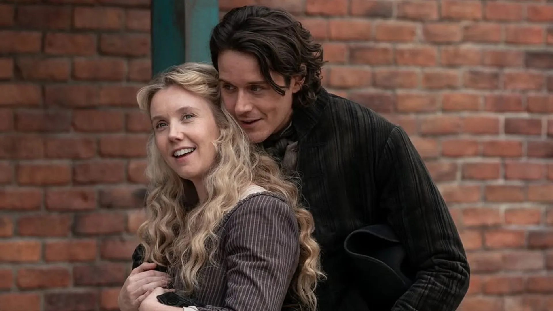 Two people in period costumes stand close together in front of a brick wall; the woman with long blonde hair is smiling, while the man leans in behind her, holding a black hat.