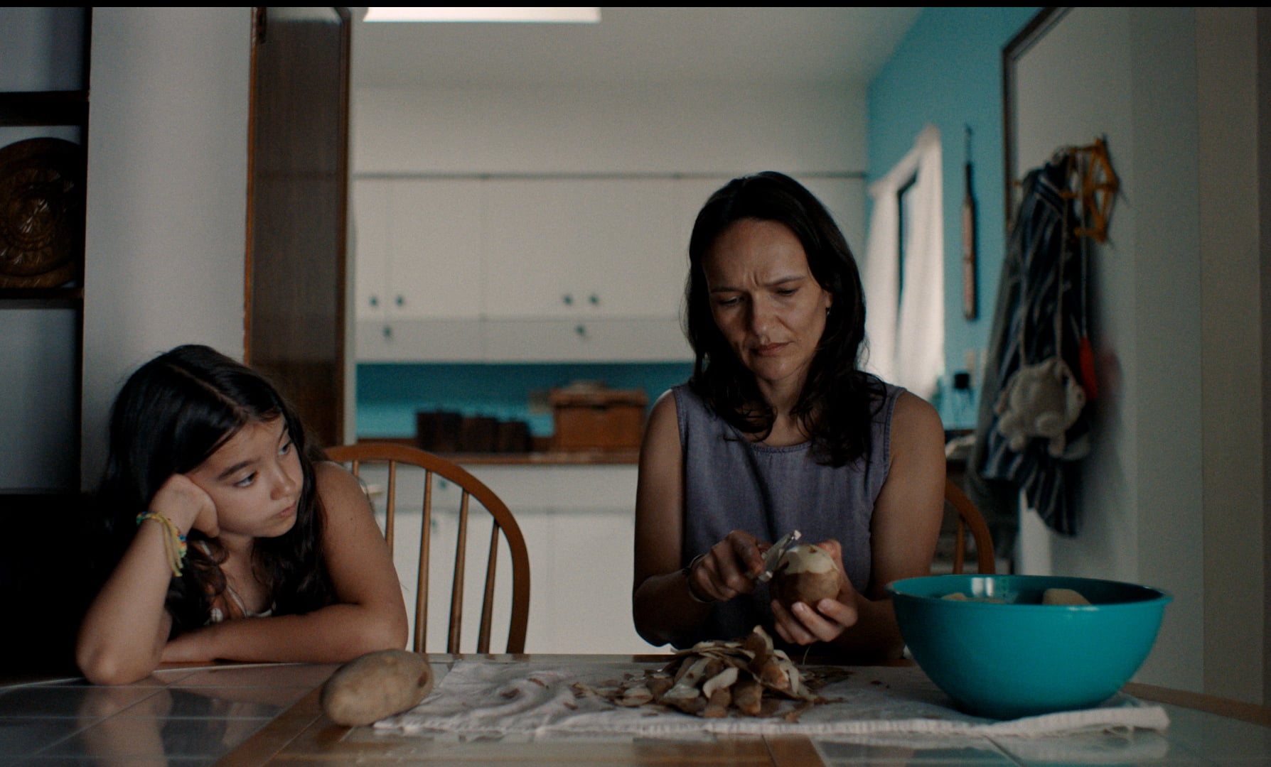 A woman peels potatoes at a kitchen table while a young girl sits beside her, resting her head on her hand and looking bored.