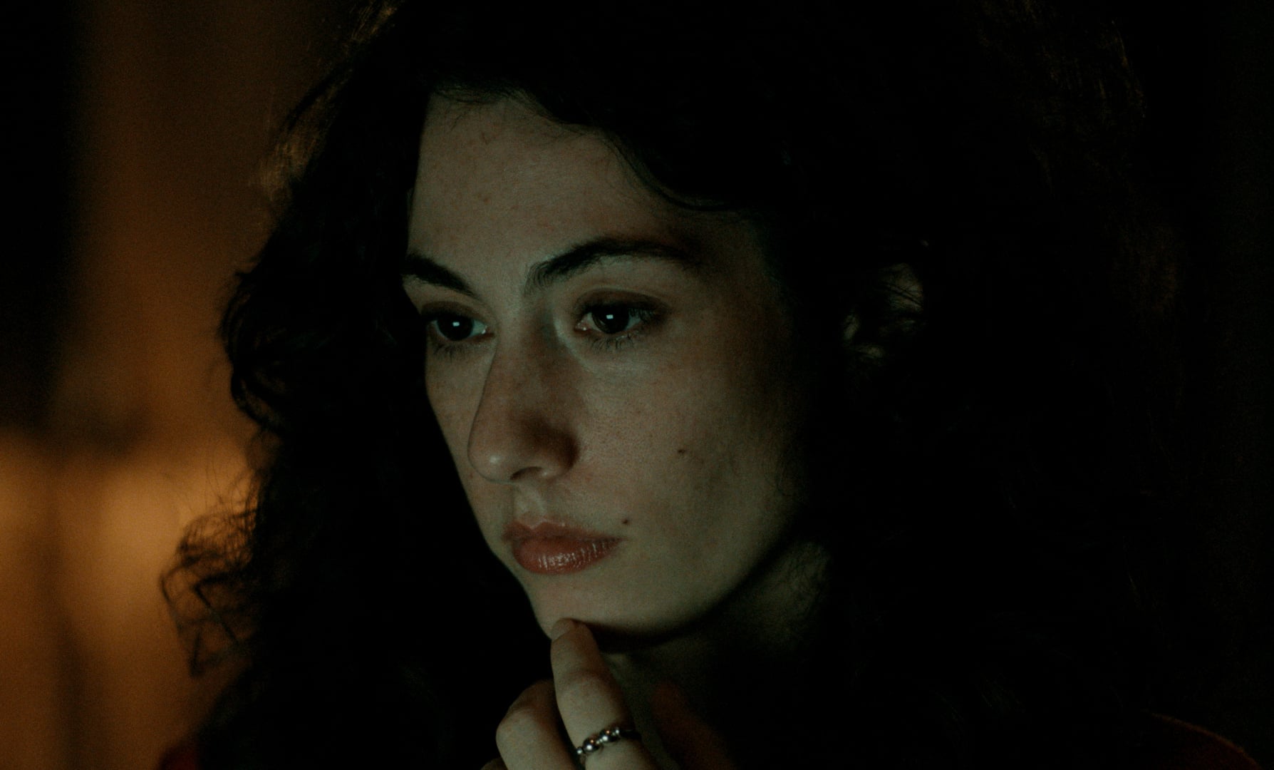 A woman with curly dark hair and a contemplative expression rests her chin on her hand, looking slightly downward in dim lighting.
