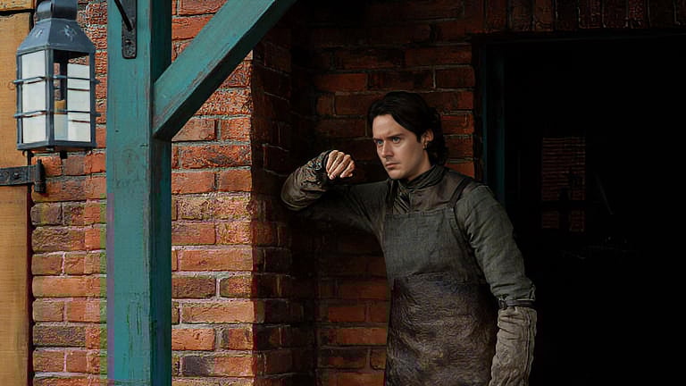 A man in medieval clothing leans against a brick wall near a wooden beam and lantern, looking pensively into the distance.
