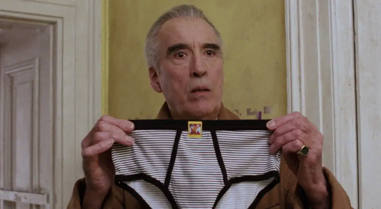 An older man with gray hair holds up a pair of black-and-white striped briefs in front of a yellow wall, looking forward with a neutral expression.