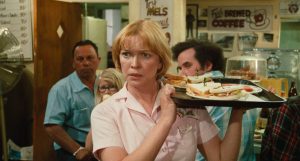 A waitress in a pink uniform holds a tray with sandwiches in a busy diner, with customers and menu signs visible in the background.