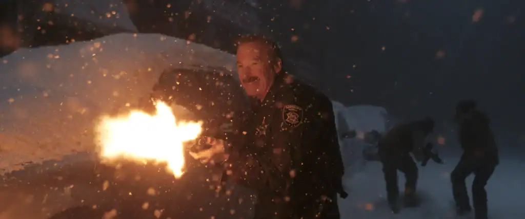 A police officer fires a gun at night in heavy snow next to a snow-covered vehicle, with two people in the background.