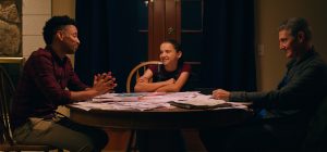 Mysterious Family Drama ‘Captain Tsunami’ Debuts Trailer Ahead of Release (EXCLUSIVE) Three people sit around a table covered with papers, engaged in conversation in a warmly lit room.