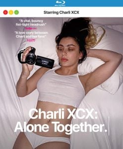 Charli XCX lies on a bed in a white outfit, holding a video camera, promoting the film "Charli XCX: Alone Together" with review quotes and Blu-ray branding.