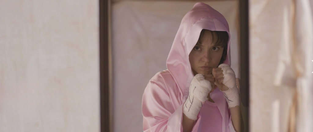 A woman wearing a pink hooded robe and hand wraps stands in a boxing stance, looking determined.