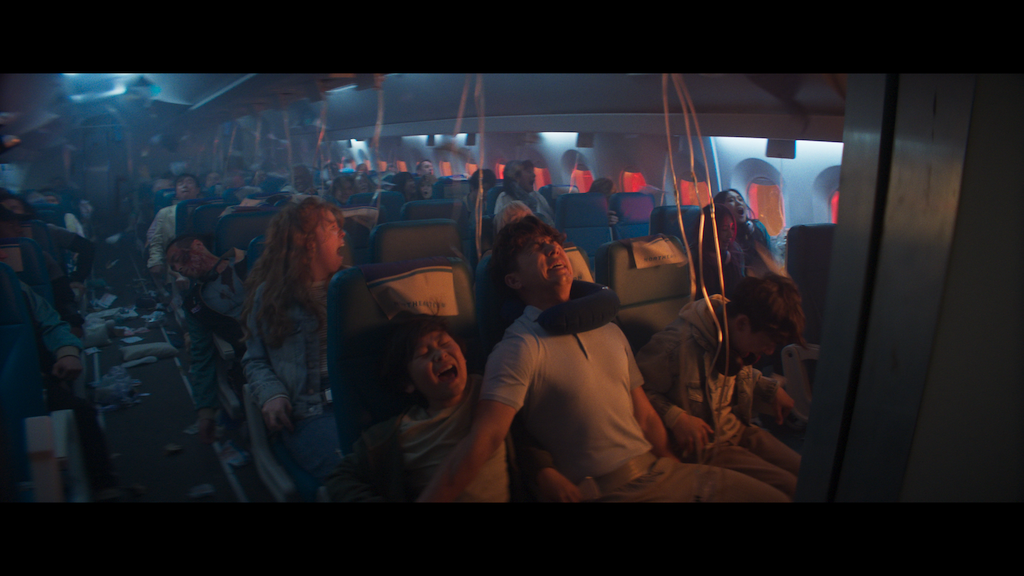 Passengers in an airplane cabin panic as oxygen masks drop and the cabin is filled with smoke and debris, indicating an emergency situation.