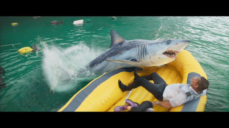 A large shark lunges out of the water toward a man in a yellow inflatable raft, splashing water and causing visible distress.