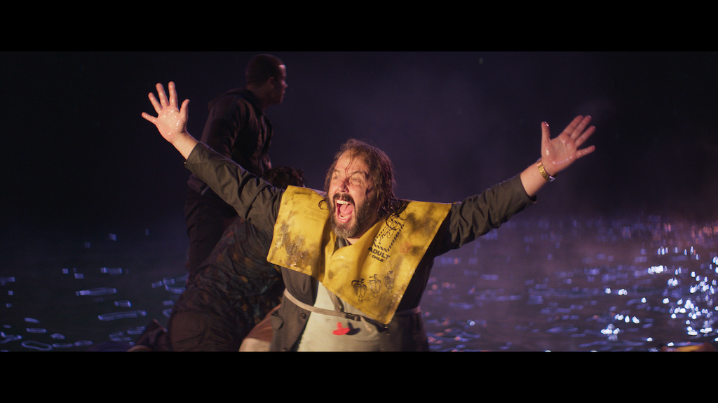 A man wearing a yellow life vest stands in waist-deep water at night, arms raised and mouth open, with two other people behind him.