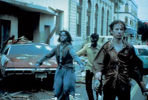 Three people dressed as zombies walk down a city street lined with debris and damaged cars, re-creating a scene from an urban disaster or apocalypse.