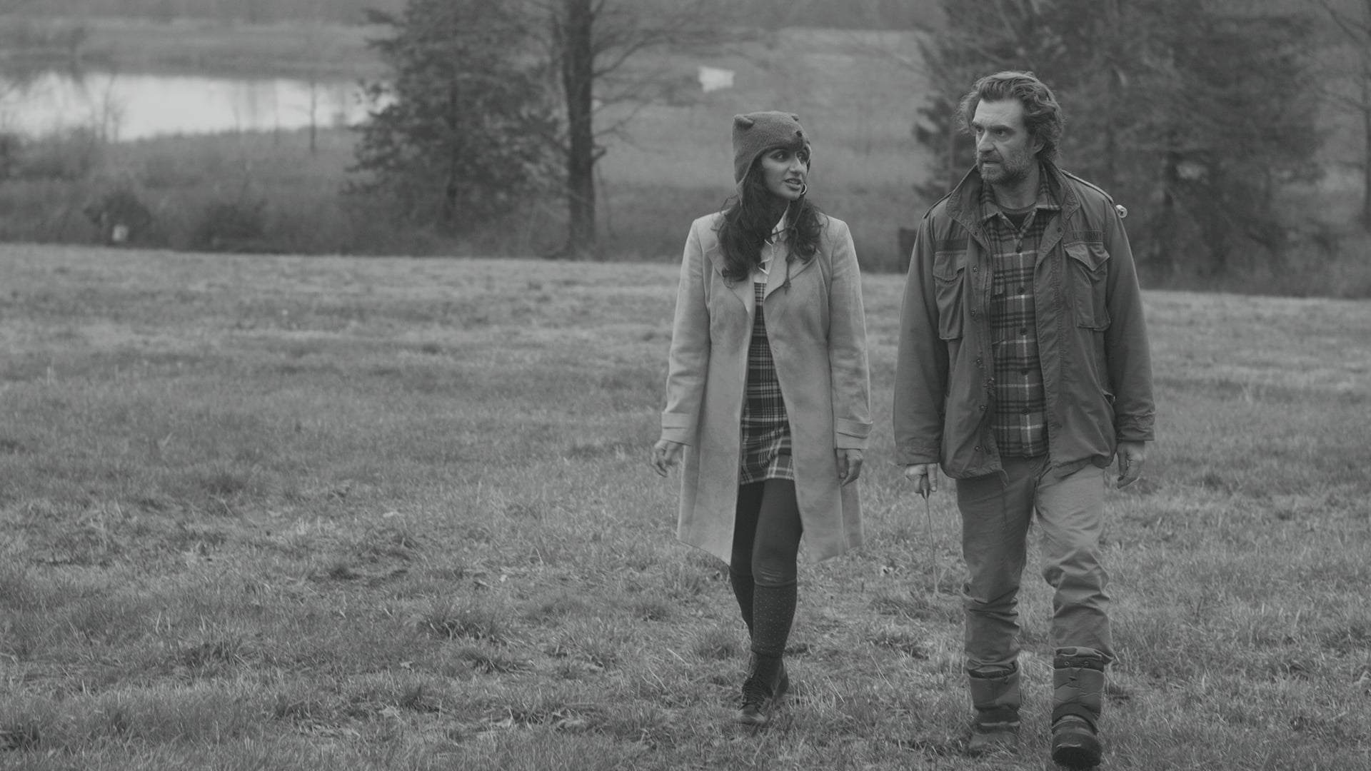 A woman and a man walk side by side across a grassy field, dressed in coats and autumn clothing, with trees and a body of water in the background.